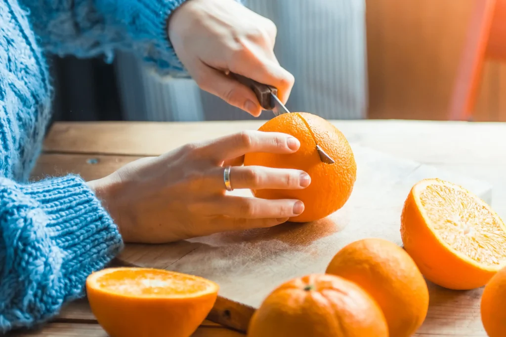 Imagem de uma mulher com suéter azul cortando uma laranja ao meio em cima de uma mesa de madeira, com outras laranjas ao redor.